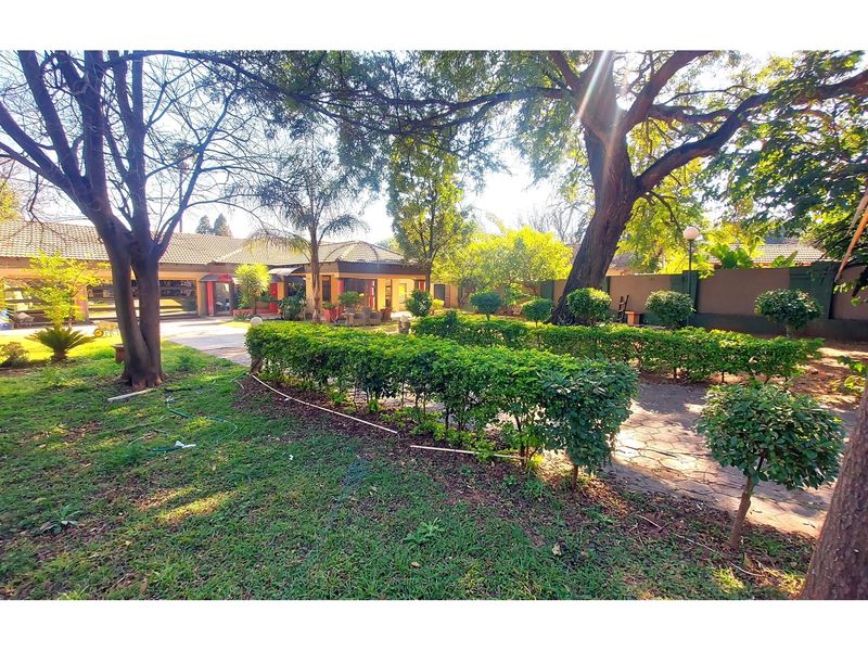 A spacious garden area featuring mature trees, manicured hedging, and a paved walkway leading towards the main house structure.