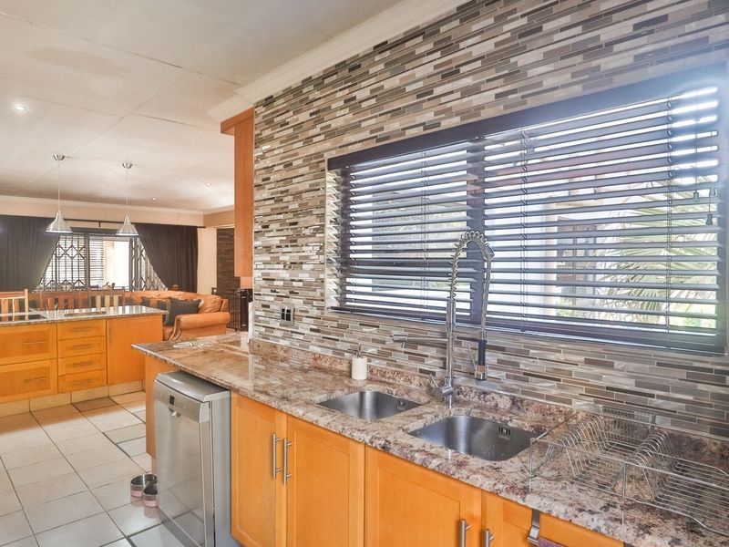 Open plan kitchen featuring granite countertops, mosaic tile backsplash, double basin sink, and warm-toned wooden cabinetry, flowing into the adjacent living area.