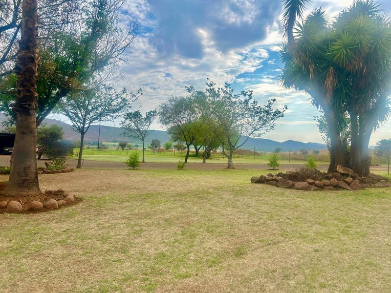 A spacious farm garden featuring mature trees, rock-bordered garden beds, and expansive views of the surrounding landscape and distant mountains.
