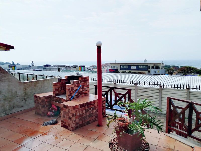 An open balcony featuring a built-in brick braai, tiled flooring, and a wooden balustrade, offering an elevated outdoor space with coastal views.