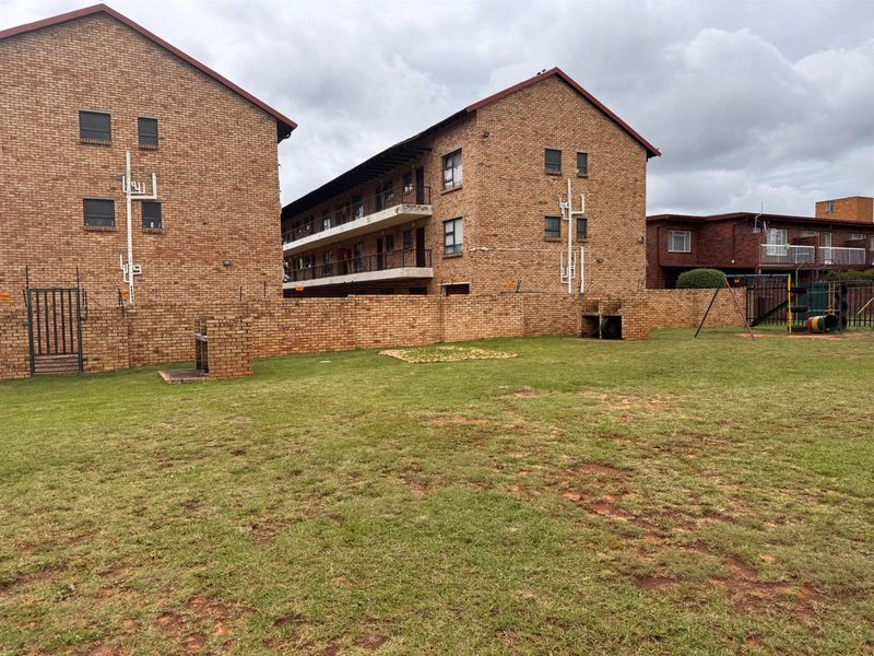 Rear view of the apartment complex featuring a communal garden area with brick boundary walls, built-in braai facilities, and a small playground.