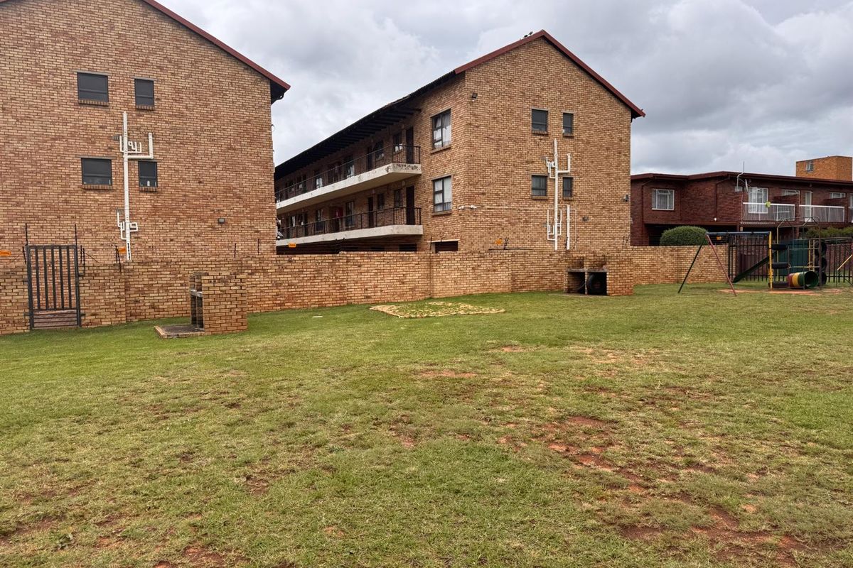 Rear view of the apartment complex featuring a communal garden area with brick boundary walls, built-in braai facilities, and a small playground.