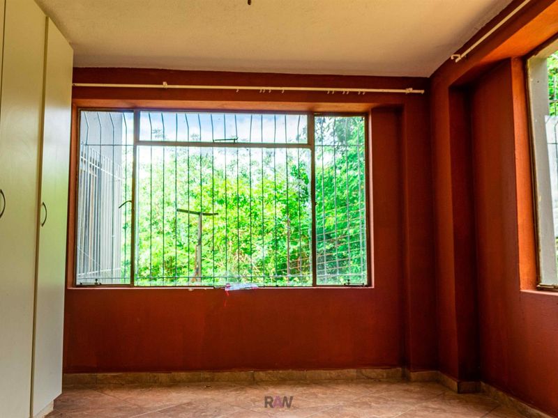 A bedroom featuring built-in cupboards, tiled flooring, and large windows with security bars overlooking greenery.