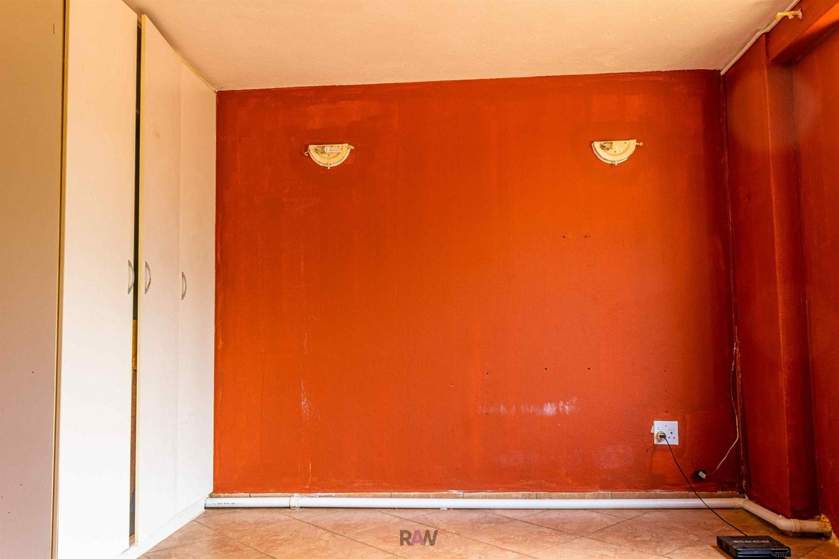 A bedroom featuring built-in cupboards, tiled flooring, and wall-mounted lighting fixtures, currently presenting with dated finishes and visible surface-mounted electrical and plumbing elements.