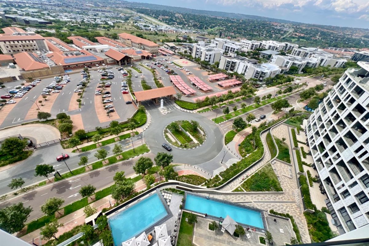 Elevated aerial view overlooking the apartment complex, featuring swimming pools, landscaped grounds, and surrounding urban infrastructure.