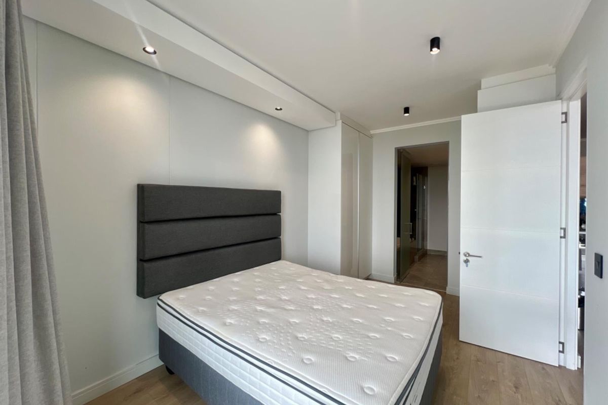 A modern bedroom featuring built-in wardrobes, recessed ceiling lighting, and light wood-look flooring, leading into an adjacent bathroom area.