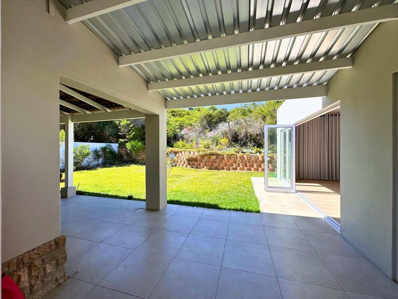 A spacious covered patio featuring tiled flooring, stone accents, and a corrugated metal roof, seamlessly connecting to the garden through large stacking glass doors.