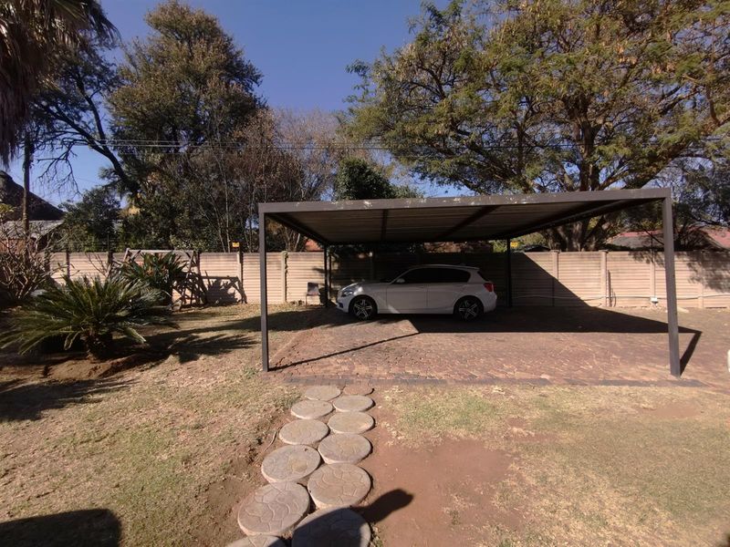 Outdoor area featuring a paved driveway, a metal carport structure, and a garden with established trees and a stone pathway.