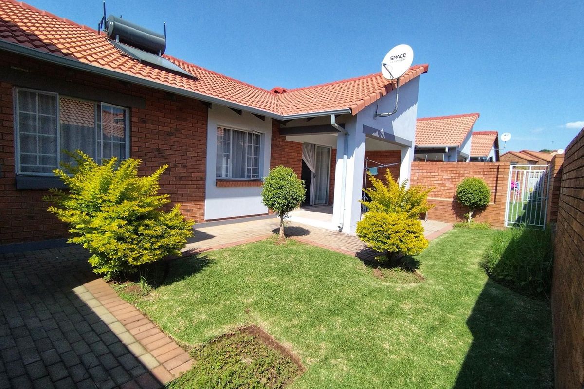 Rear view of a townhouse featuring a brick facade, tiled roof with solar water heater, paved patio area, and a private garden with boundary wall.