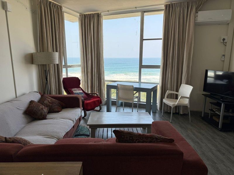 A living room featuring large floor-to-ceiling windows with direct ocean views, laminate flooring, and a wall-mounted air conditioning unit.