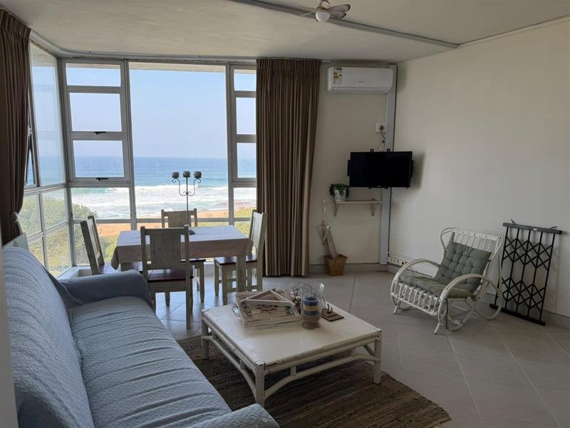 Open-plan living and dining area featuring large windows with direct ocean views, tiled flooring, and a wall-mounted air conditioning unit.