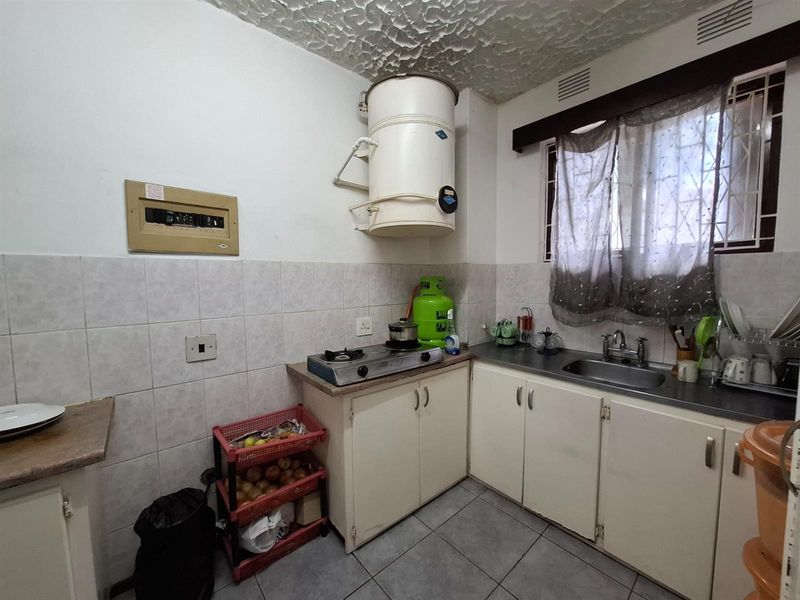 A compact kitchen featuring tiled flooring, built-in cabinetry, a stainless steel sink, and a wall-mounted geyser, offering a functional space with potential for modernization.