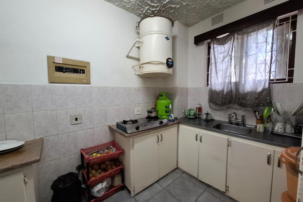 A compact kitchen featuring tiled flooring, built-in cabinetry, a stainless steel sink, and a wall-mounted geyser, offering a functional space with potential for modernization.