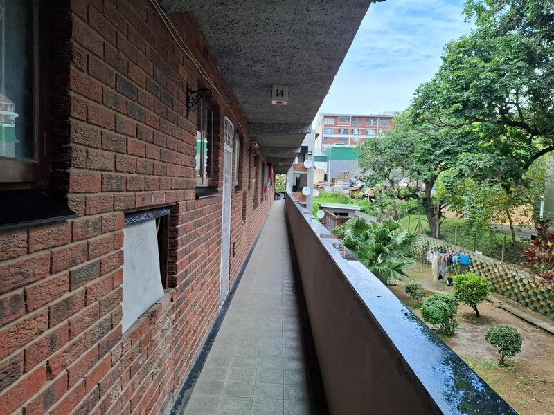 An exterior apartment walkway featuring exposed brick walls, tiled flooring, and a metal balustrade overlooking the communal garden area.