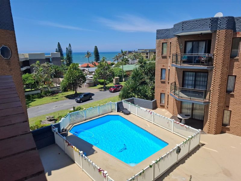 Elevated view of the apartment complex featuring a communal swimming pool with safety fencing and surrounding landscaped grounds with ocean views.