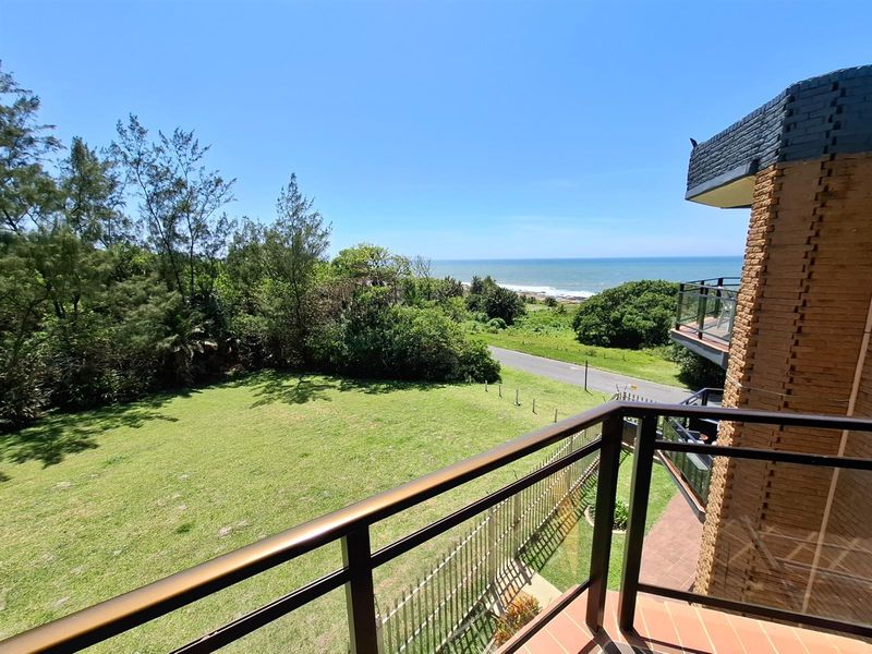 A scenic view from the balcony overlooking a lush green lawn, coastal vegetation, and the ocean in the distance.