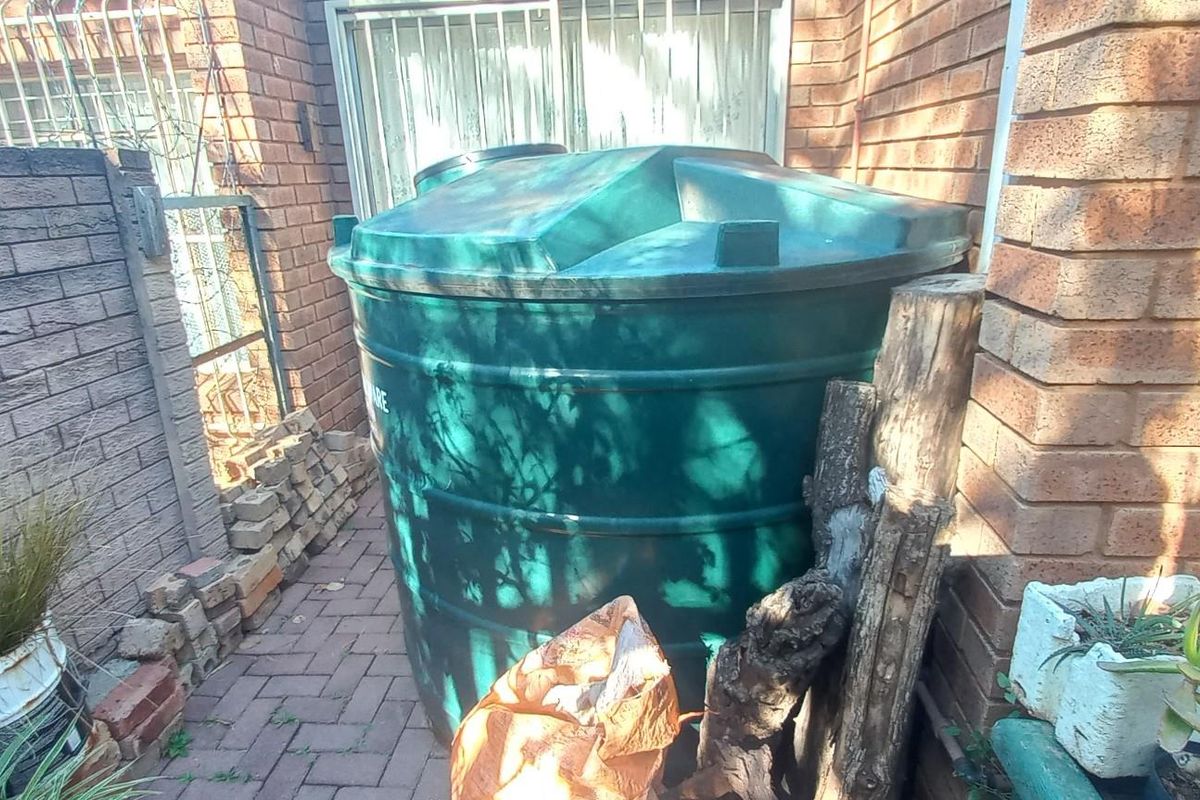 A rear exterior area featuring a large green water storage tank positioned against a brick wall with paved ground.