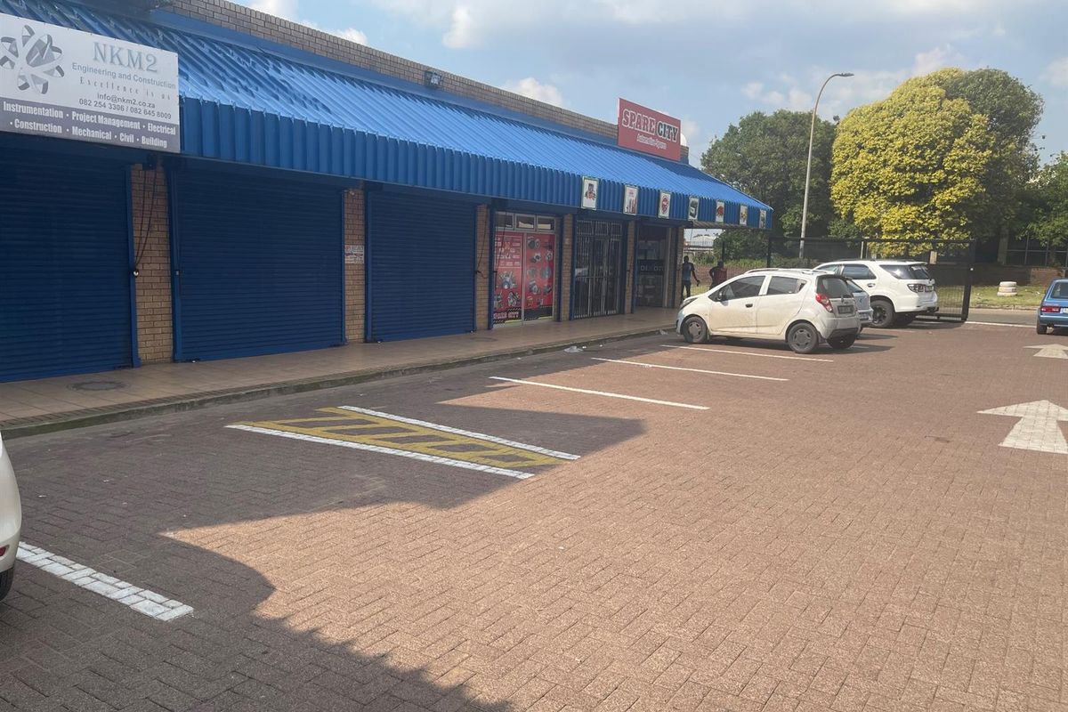 Exterior view of a retail strip center featuring blue metal awnings, roller shutter doors, and a paved parking area in front.