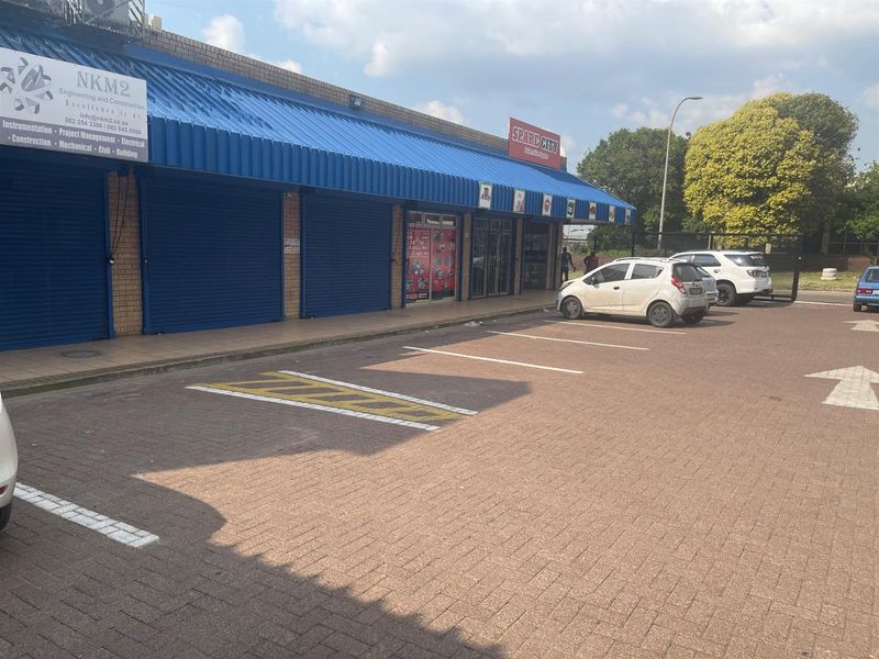 Exterior view of a retail strip center featuring blue metal awnings, roller shutter doors, and a paved parking area in front.