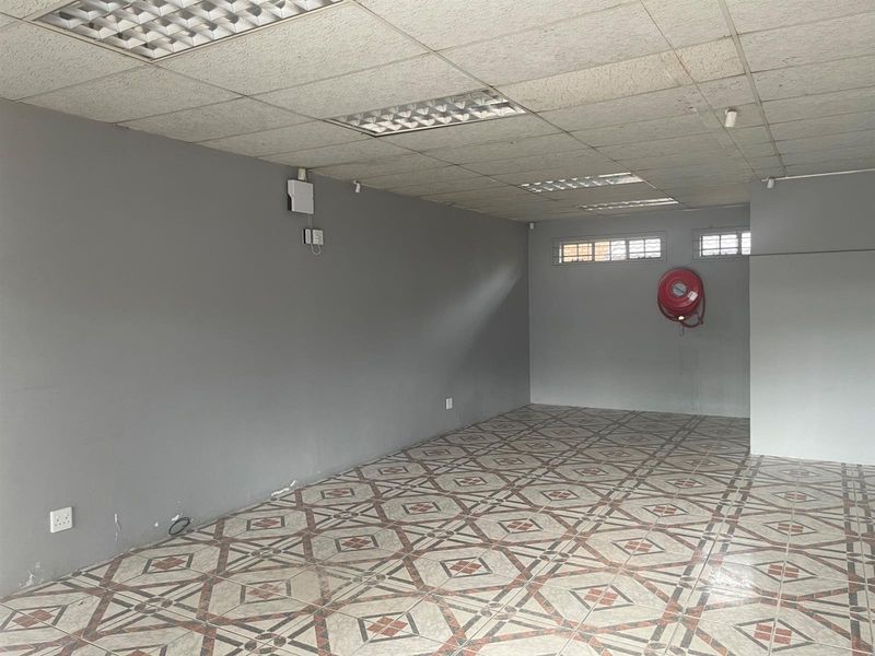 An empty retail space featuring patterned ceramic floor tiles, grey painted walls, recessed fluorescent lighting, and a wall-mounted fire hose reel.