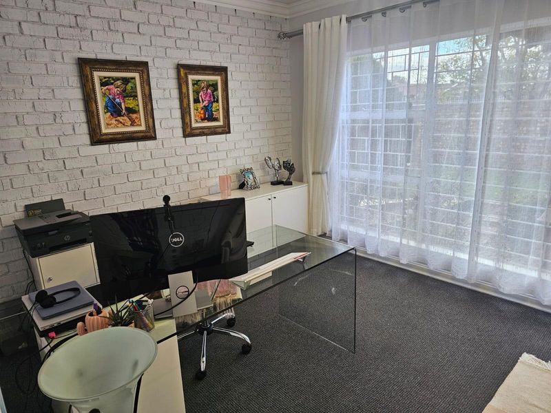 A functional home office featuring a textured white brick feature wall, neutral carpeting, and large windows with security bars.