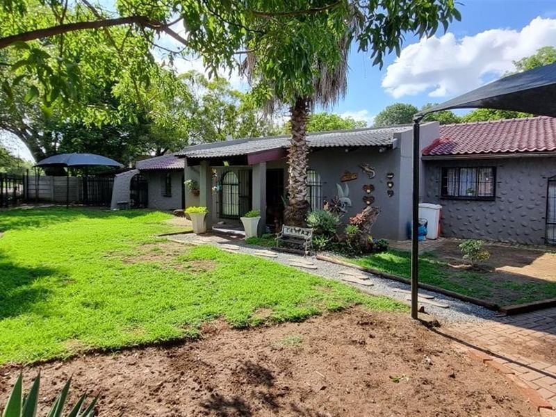Rear view of the property featuring a landscaped garden, paved driveway area, and covered patio space with security fixtures.