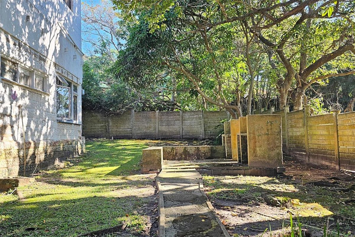 A rear garden area featuring a concrete pathway, perimeter boundary walls, and a built-in masonry barbecue structure in need of maintenance.