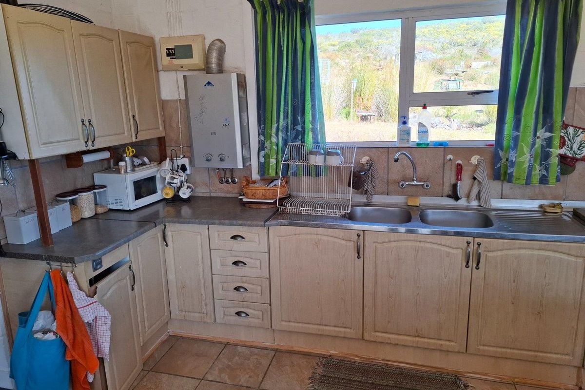A functional kitchen featuring light wood cabinetry, a double stainless steel sink, tiled flooring, and a wall-mounted gas geyser with a view of the landscape.