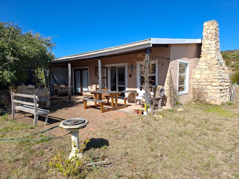 A rustic farm cottage featuring a covered patio area, stone chimney, and sliding glass doors, set within a natural landscape.