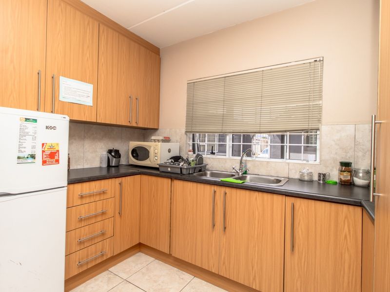 A functional office kitchen featuring light wood-finish cabinetry, dark laminate countertops, a double basin sink, and tiled flooring with a window for natural light.
