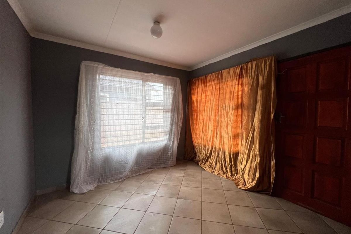 A simple bedroom featuring tiled flooring, a large window with security bars, and a solid timber door.