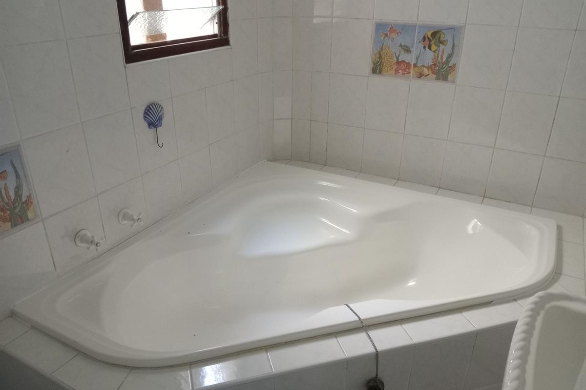A corner bathtub featuring white tiled surround and decorative accent tiles, set against a window with louvered glass.