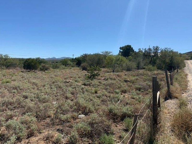 A view of a vacant residential plot featuring natural vegetation and a wire fence boundary along the roadside.