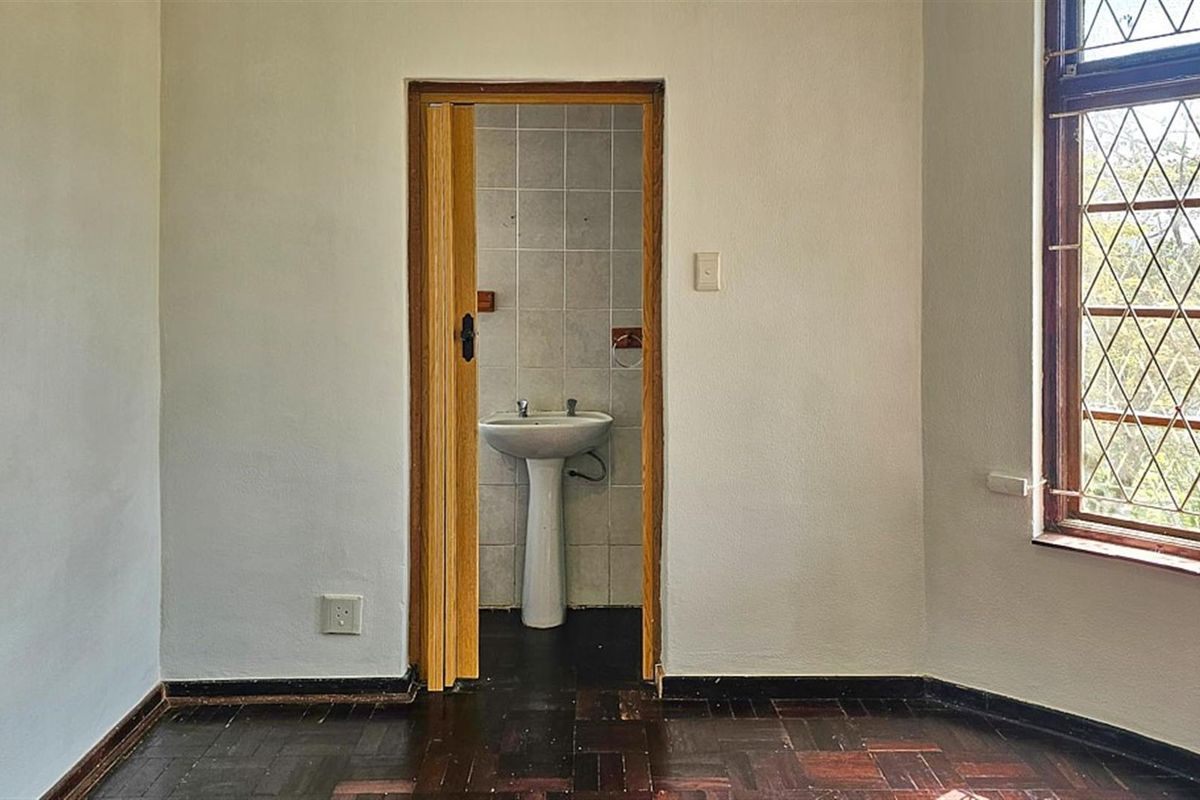 A view from a room with dark parquet flooring looking into a small, dated bathroom featuring a pedestal basin and tiled walls.