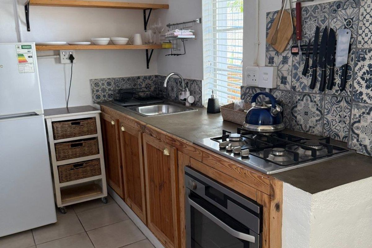 Compact kitchen featuring wooden cabinetry, built-in oven, gas hob, stainless steel sink, and decorative patterned tile backsplash with open shelving.