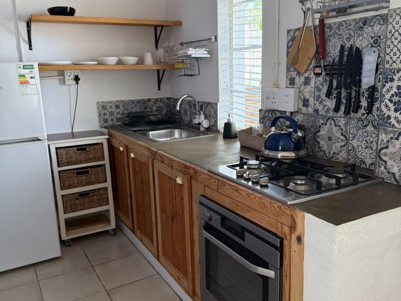 Compact kitchen featuring wooden cabinetry, built-in oven, gas hob, stainless steel sink, and decorative patterned tile backsplash with open shelving.