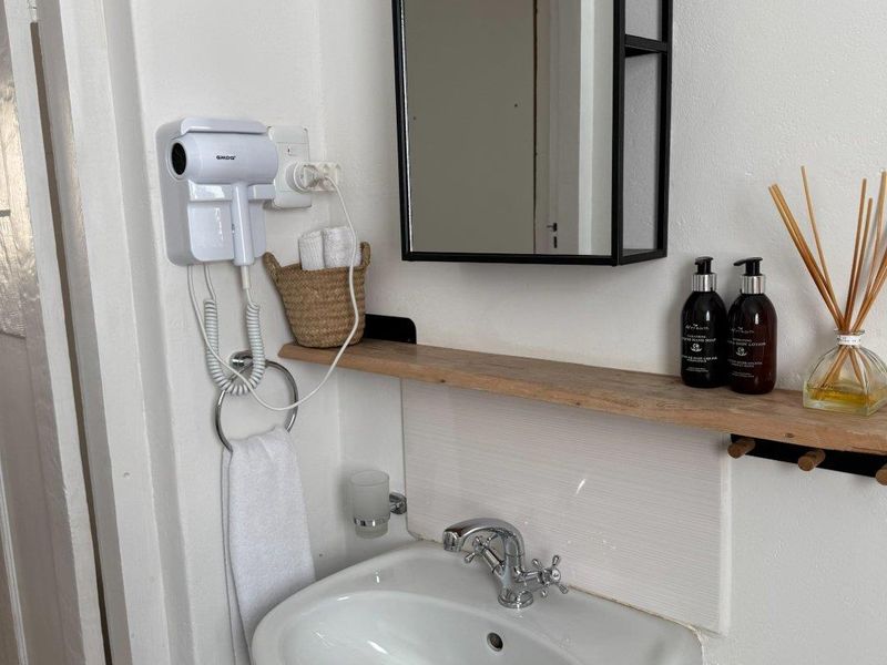 A clean and functional bathroom vanity area featuring a pedestal sink, chrome faucet, wall-mounted mirror, and convenient wooden shelving.