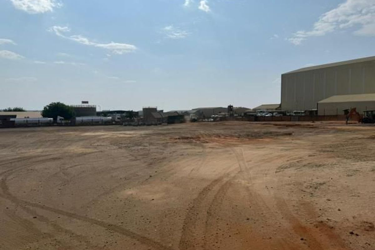 Expansive industrial yard area with unpaved ground, featuring surrounding warehouse structures and storage tanks in an industrial estate setting.