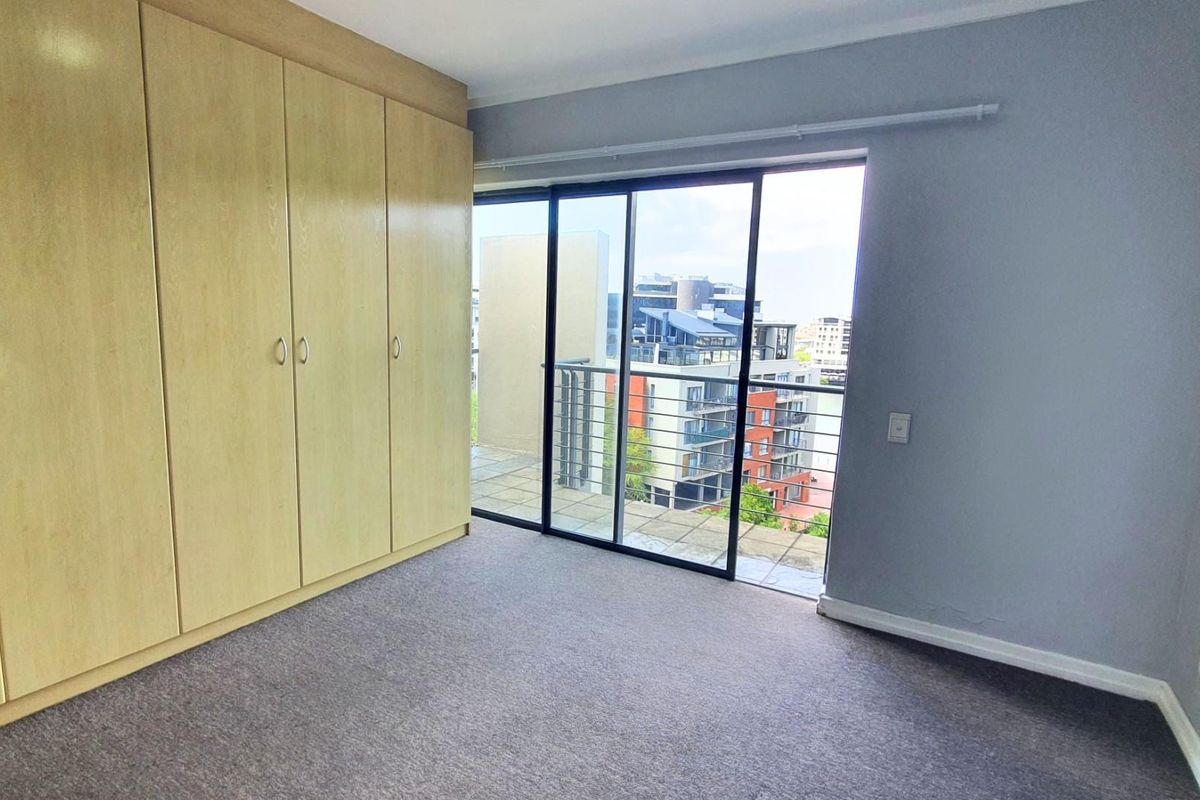 A bright bedroom featuring built-in wooden cabinetry, neutral carpeting, and large sliding glass doors opening onto a private balcony with urban views.