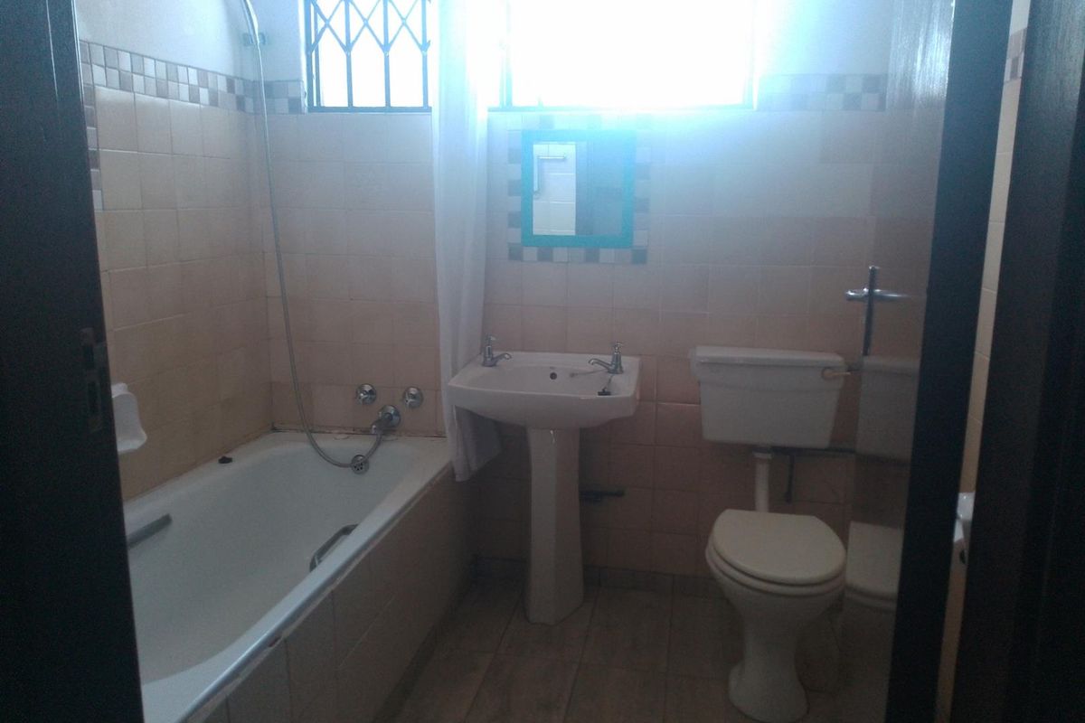 A functional bathroom featuring a pedestal basin, toilet, and bathtub with shower mixer, finished with dated wall tiling and a security-fitted window.