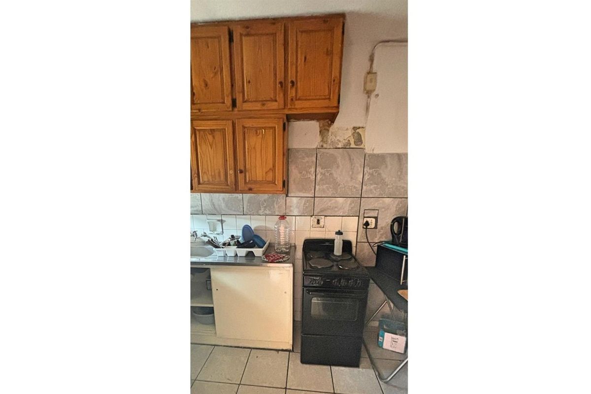 A dated kitchen area featuring wooden wall-mounted cabinetry, tiled splashbacks, and a freestanding electric stove, requiring significant renovation and repair.