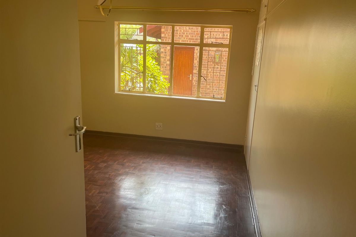 A vacant bedroom featuring classic parquet flooring, built-in cupboards, and a large window providing natural light and a view of the exterior brickwork.