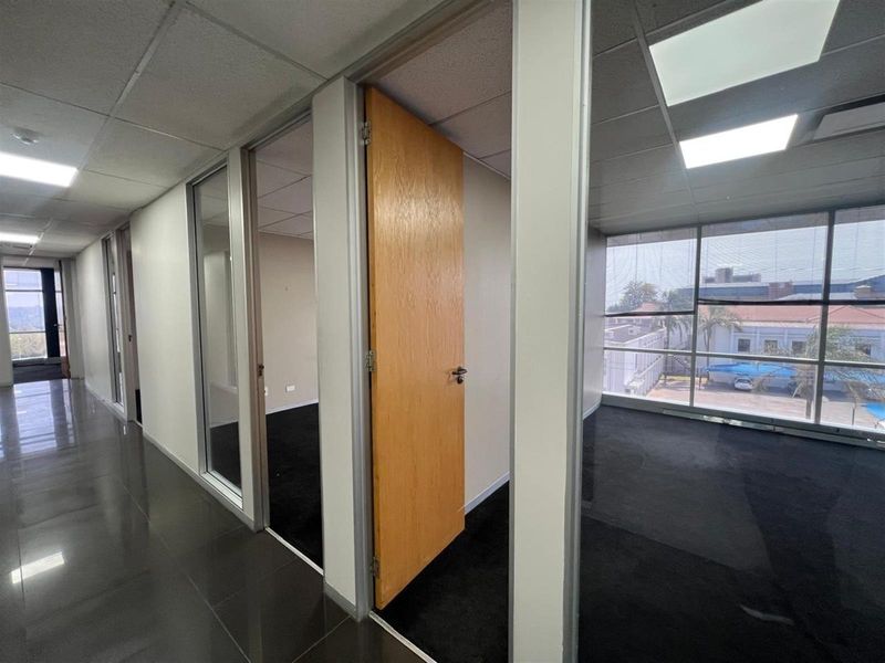 A view of an office hallway featuring glass partitions, wooden doors, and large windows providing natural light and exterior views.