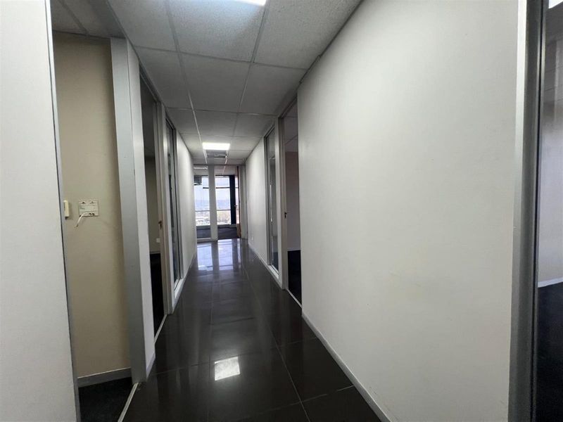 A functional office hallway featuring tiled flooring, drop ceiling panels, and multiple glass-paneled office entryways leading to a bright windowed end.