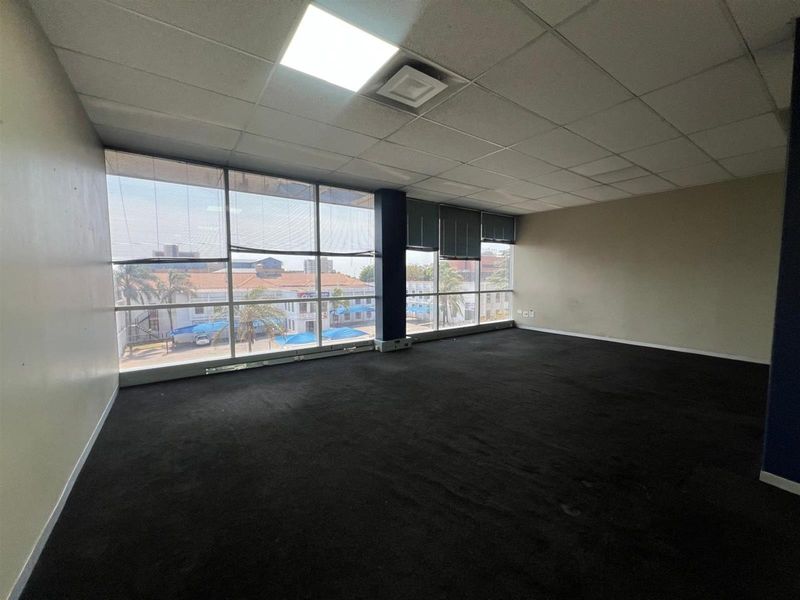 A spacious, empty office suite featuring large windows with natural light, suspended ceiling tiles, and dark carpeting, suitable for a professional workspace.
