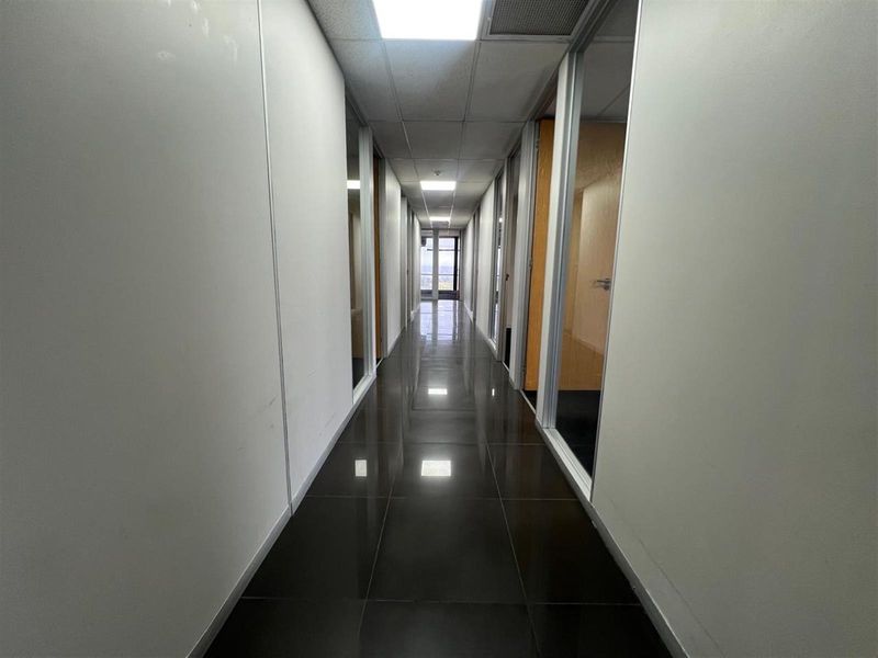 A long office corridor featuring polished dark floor tiles, glass-partitioned office doors, and a suspended acoustic ceiling with integrated lighting.