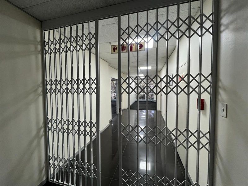 A secure office hallway featuring a retractable metal security gate, tiled flooring, and suspended ceiling tiles with integrated fire safety signage.