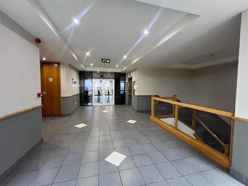 A well-maintained office building lobby featuring tiled flooring, recessed ceiling lighting, a wooden staircase railing, and glass-paneled double doors leading to the exterior.