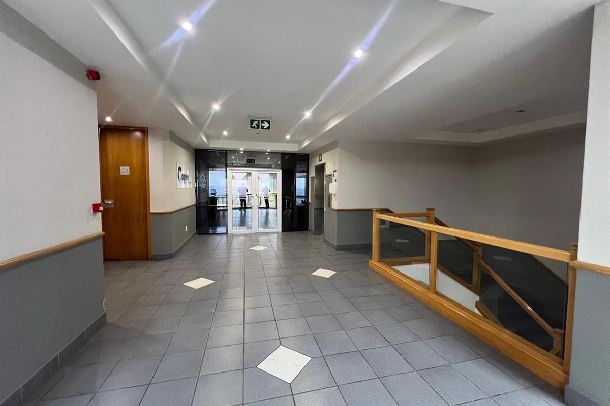 A well-maintained office building lobby featuring tiled flooring, recessed ceiling lighting, a wooden staircase railing, and glass-paneled double doors leading to the exterior.