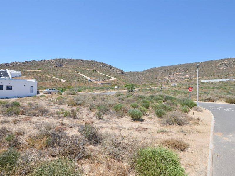 A view of a vacant residential plot featuring natural vegetation, adjacent to a paved road and existing residential infrastructure.